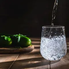 Woda nalewana do szklanki, obok leżą ogórki na desce.