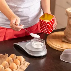 Osoba odmierzająca cukier łyżką na wagę kuchenną, obok jajka i mąka w worku.