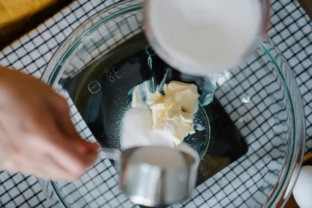 Kobieta wsypuje cukier do miski z masłem na wadze kuchennej.